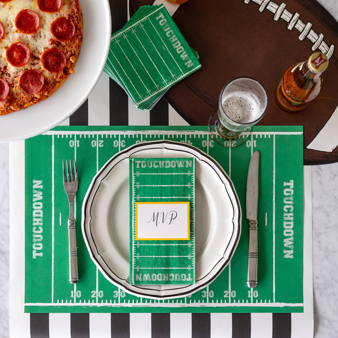 A football-themed place setting with football field styled placemats, napkins, and name card, football shaped placemat, beer, black and white striped table runner, and pizza.