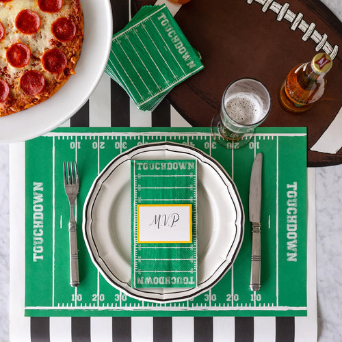 A football-themed place setting with football field styled placemats, napkins, and name card, football shaped placemat, beer, black and white striped table runner, and pizza.