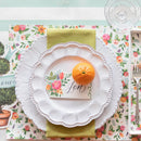 Table setting with floral placemat, white plate, orange, and decorative card.
