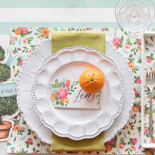 Table setting with floral placemat, white plate, orange, and decorative card.