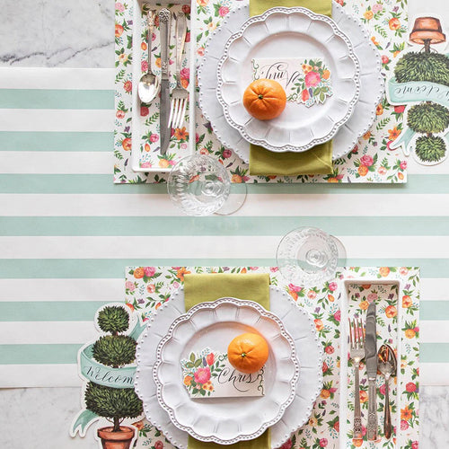 Table setting with floral placemats, white plates, and cutlery on a striped tablecloth.