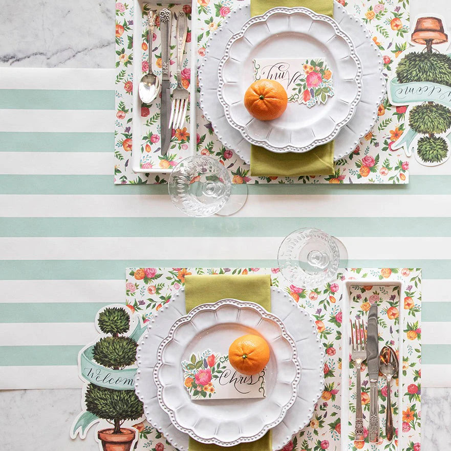 Table setting with floral placemats, white plates, and cutlery on a striped tablecloth.
