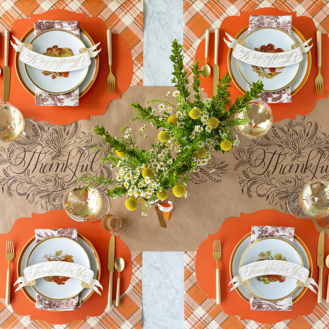 A table set for Thanksgiving with a brown table runner with "thankful" in script, flowers in the center, and four place settings with orange accents and place cards shaped like banners with 'thankful for you' written on them.