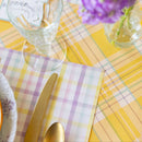 Table setting with yellow and purple checkered tablecloth, gold cutlery, and a vase with purple flowers.