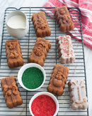 Molded cakes featuring gifts, snowmen, and other Christmas themed shapes on a cooking rack with containers of icing and sprinkles.