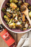 Bowl of roasted brussels sprouts with a wooden spoon, next to a bottle of hot honey on a wooden surface.