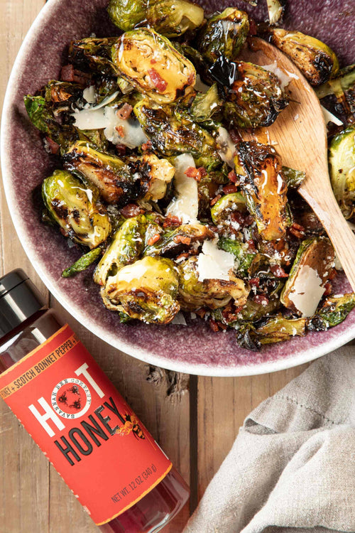 Bowl of roasted brussels sprouts with a wooden spoon, next to a bottle of hot honey on a wooden surface.