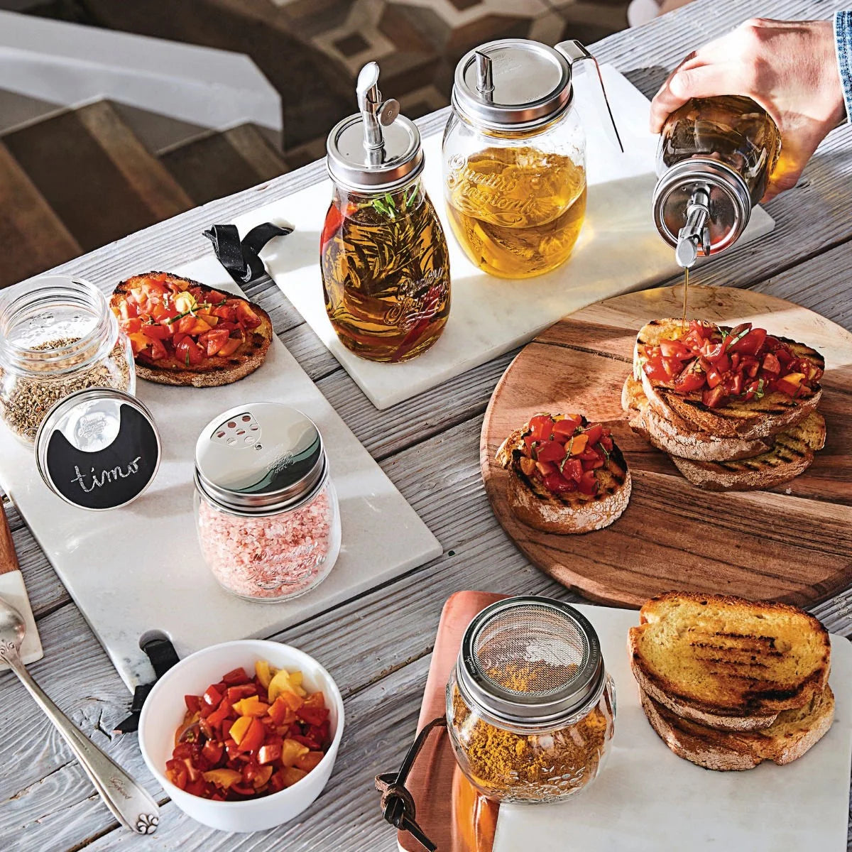 A styled tabletop scene with glass mason jars containing infused oils and spices, alongside bowls of grilled toast topped with diced tomatoes and herbs. A hand is visible pouring oil from one of the jars, and the setting includes a wooden cutting board, white platter, and rustic wood table.