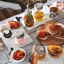 A styled tabletop scene with glass mason jars containing infused oils and spices, alongside bowls of grilled toast topped with diced tomatoes and herbs. A hand is visible pouring oil from one of the jars, and the setting includes a wooden cutting board, white platter, and rustic wood table.