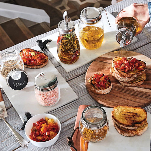 A styled tabletop scene with glass mason jars containing infused oils and spices, alongside bowls of grilled toast topped with diced tomatoes and herbs. A hand is visible pouring oil from one of the jars, and the setting includes a wooden cutting board, white platter, and rustic wood table.