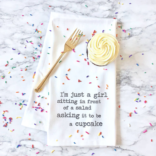 Kitchen towel with text on a marble surface with a gold fork, cupcake, and sprinkles.