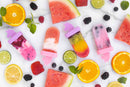 A collection of ice pops with unicorn shaped handles on a marble surface with berries and watermelon slices.