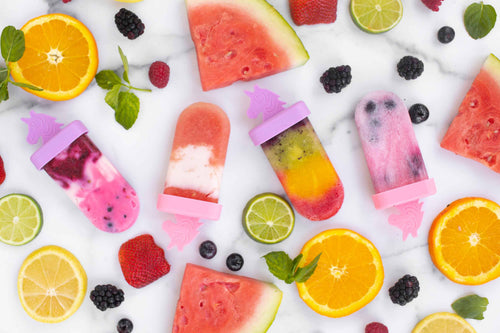 A collection of ice pops with unicorn shaped handles on a marble surface with berries and watermelon slices.