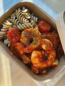 A close-up overhead shot of a meal prep container filled with pasta, shrimp, halved cherry tomatoes, and garlic in a rich red sauce, garnished with herbs.