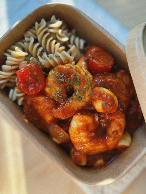A close-up overhead shot of a meal prep container filled with pasta, shrimp, halved cherry tomatoes, and garlic in a rich red sauce, garnished with herbs.