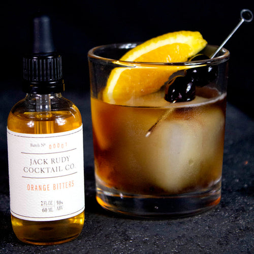 A bottle of Jack Rudy Orange Bitters beside a cocktail on a dark surface.