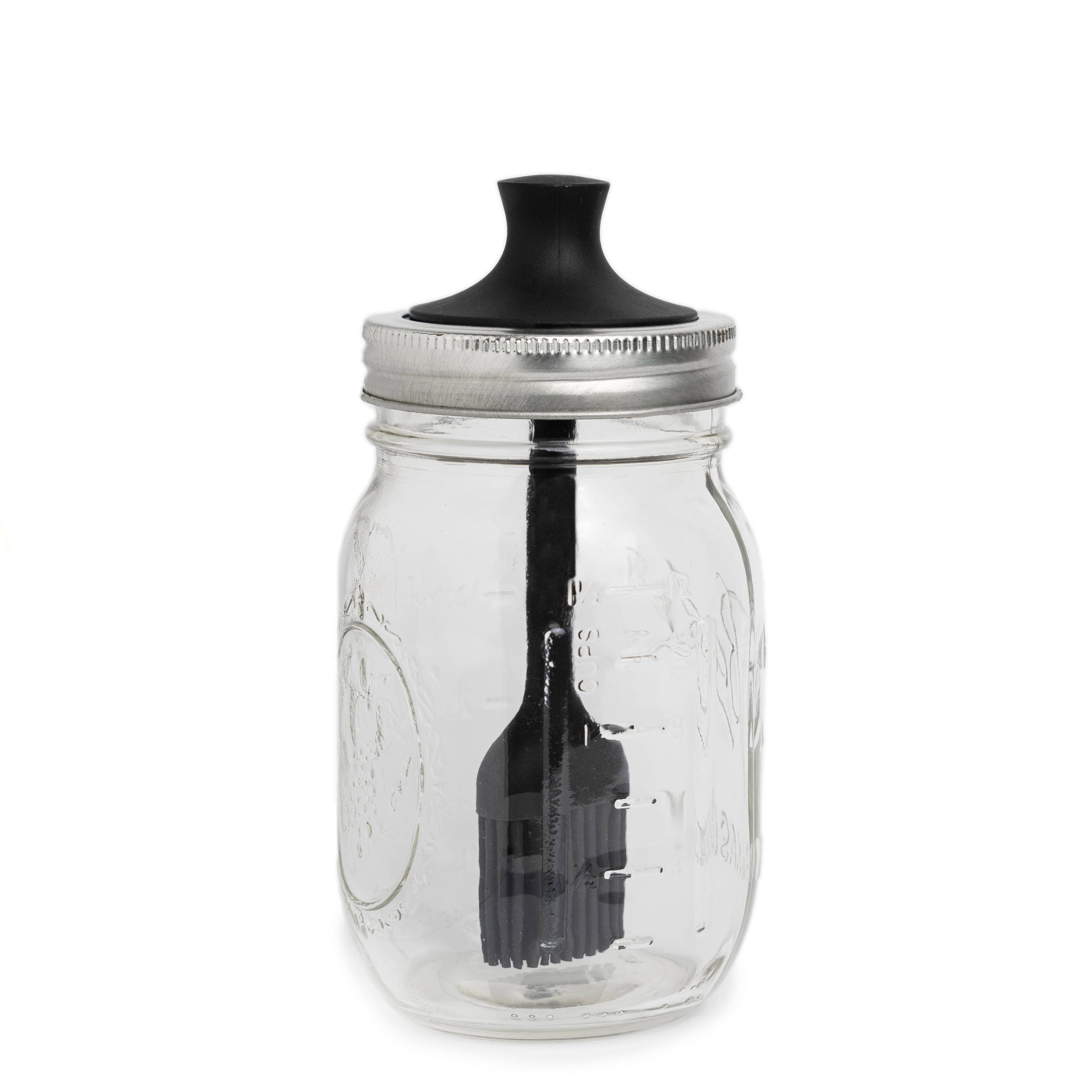 Clear glass mason jar with a black lid and attached silicone brush on a white background