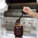 Hand using a spoon to scoop barbecue sauce from a jar on a kitchen counter.