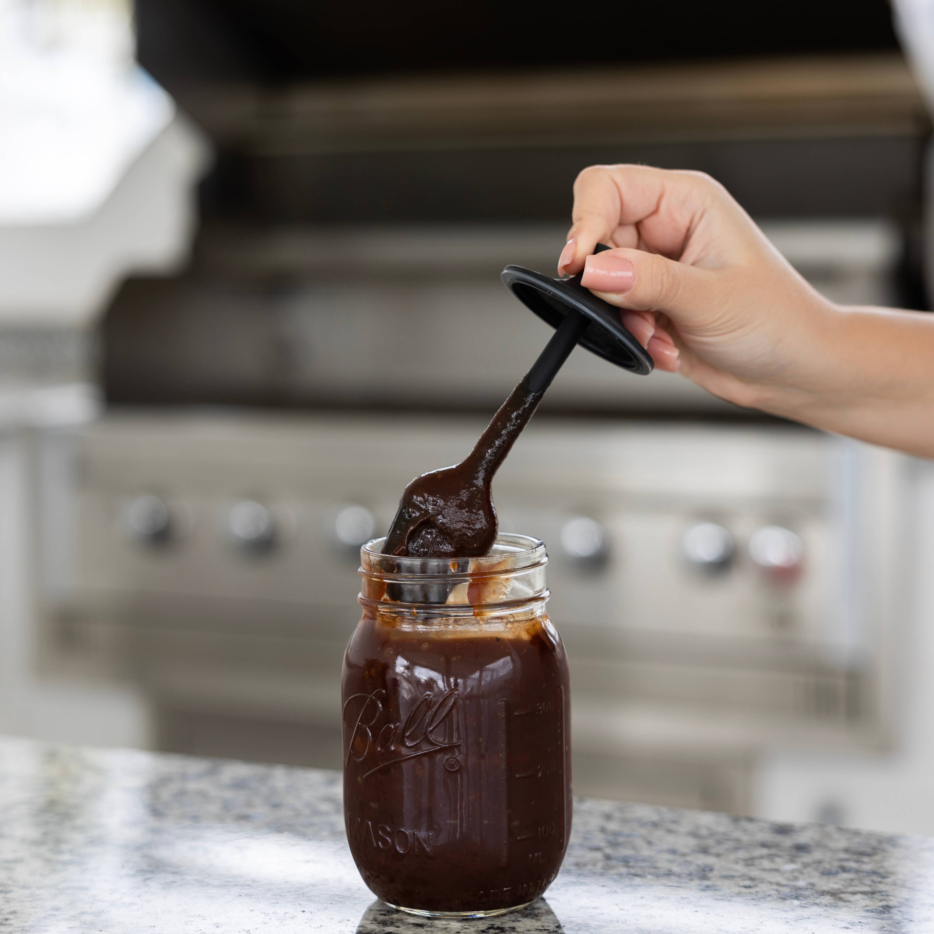 Hand using a spoon to scoop barbecue sauce from a jar on a kitchen counter.