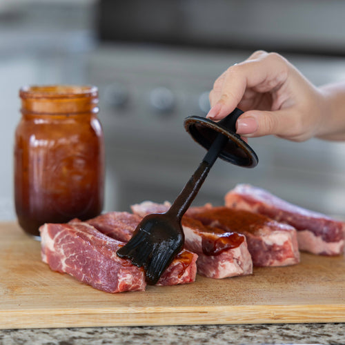 Person applying barbecue sauce to raw meat with a brush on a cutting board.
