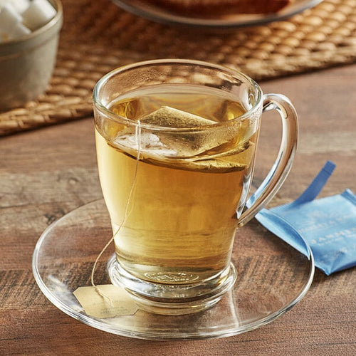 A clear glass mug of green tea on a saucer next to an open tea bag on a wooden surface.