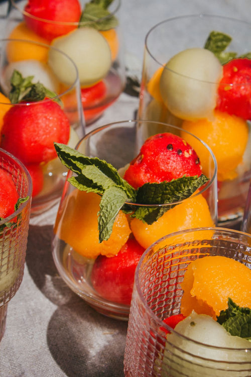 Several clear textured glass cups filled with fresh fruit, including red watermelon balls, orange melon pieces, pale cantaloupe, and mint leaves, arranged on a light gray surface.