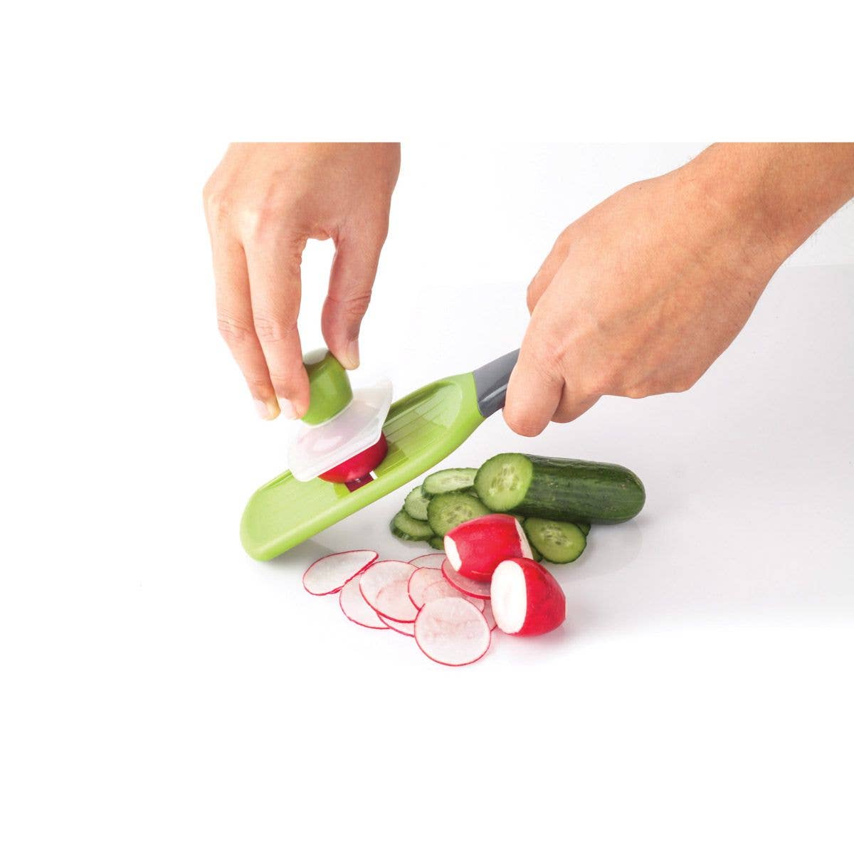 Person using a green and red vegetable slicer to cut cucumbers and radishes on a white background