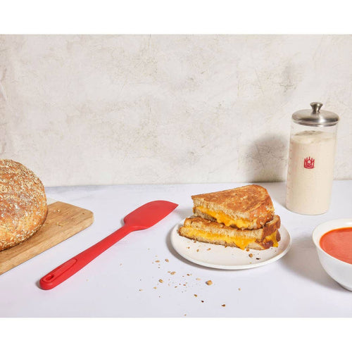Grilled cheese sandwich on a plate with a jar of sourdough starter and a bowl of soup on a white surface.