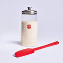 Glass container filled with sourdough with a red lid and a red spatula on a light gray background