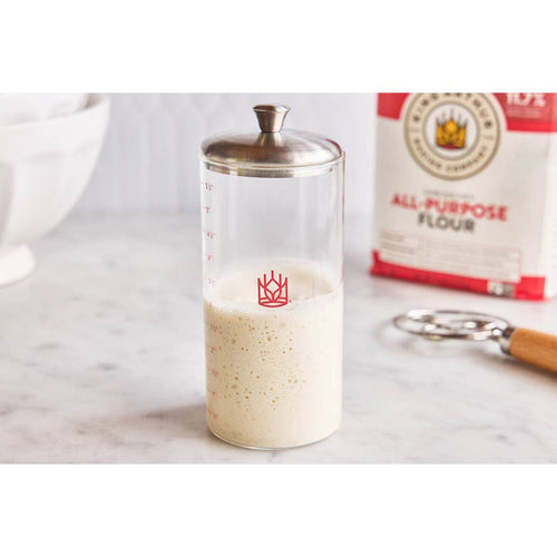 Glass container with a red logo, flour bag, and measuring spoon on a marble surface