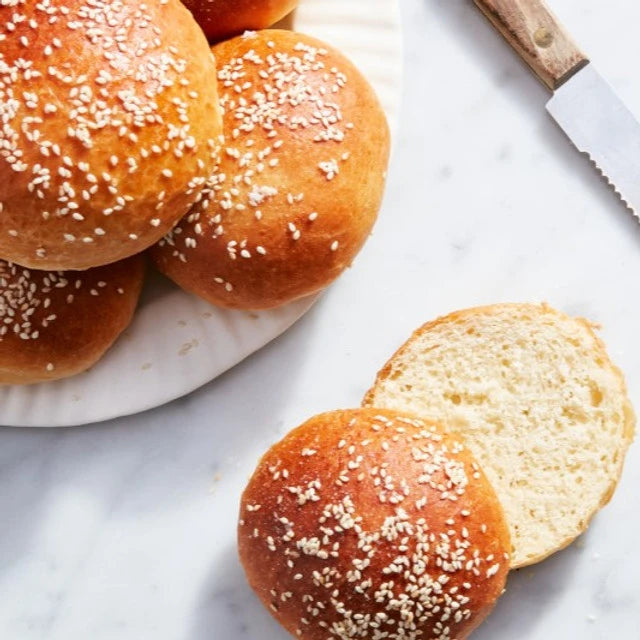 Buns with sesame seeds on a plate and one sliced open on a marble surface.