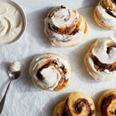 Cinnamon rolls with white icing on a white surface with a bowl of icing and a spoon.