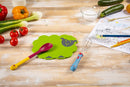 A green sheep-shaped silicone mat with a gray sheep face, shown on a light wooden table with kitchen utensils including a yellow and red spoon, a blue whisk, and a coloring page. Fresh vegetables like tomatoes, peppers, and onions are arranged around the product.