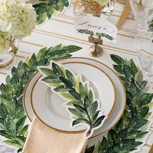 Table setting with laurel leaf-patterned placemats and cutlery on a white tablecloth.
