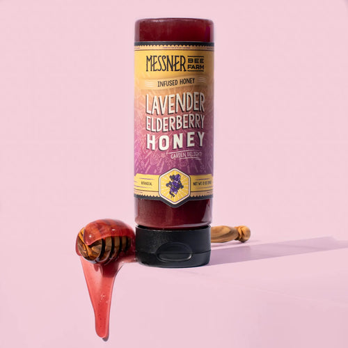 Honey container with lavender elderberry flavor with a honey dipper that is dripping honey on a pink background.