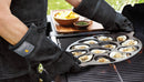 Person grilling with an oyster pan on a barbecue, wearing heat-resistant gloves.