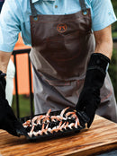 Person wearing a brown apron with a logo, holding a tray of shrimp while wearing black leather gloves on a wooden surface.