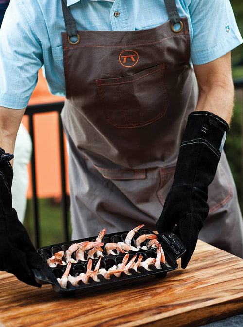 Person wearing a brown apron with a logo, holding a tray of shrimp while wearing black leather gloves on a wooden surface.