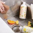 Bottle of Mandarin Coriander  and Lemon Leaf surface scrub bottles in a sink with orange and lemon slices and a person wiping down the sink's surface.