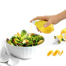 A hand holding a lemon lime mister over a bowl of salad, with lemon and lime slices visible and a pair of kitchen utensils in the background.