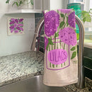 A lilac dish towel hanging on a silver faucet in a kitchen with a matching Swedish dish cloth on the wall nearby.