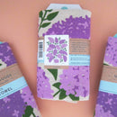 A packaged lilac dishtowel with two others on a pink background.