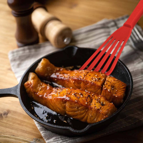 Salmon fillets in a cast iron skillet with a red fish spatula on a wooden surface.