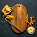 Wooden cutting and charcuterie board with a knife, yellow pepper, and small dish of spices on a dark surface