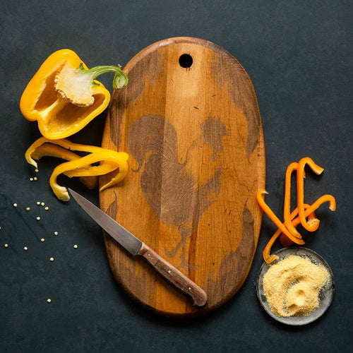 Wooden cutting and charcuterie board with a knife, yellow pepper, and small dish of spices on a dark surface