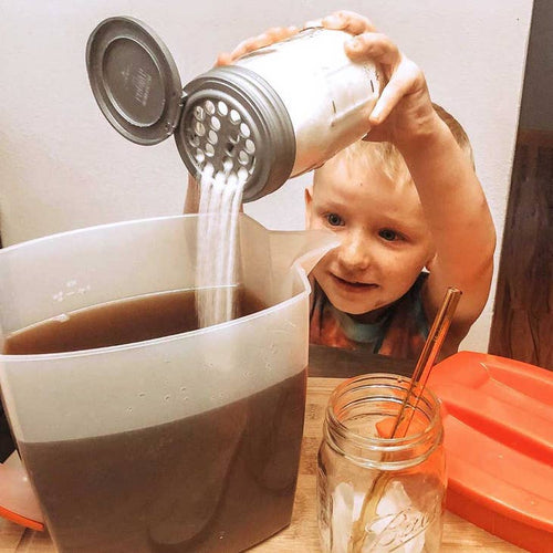 A child pouring sugar into a pitcher of liquid using a mason jar with a shaker lid.