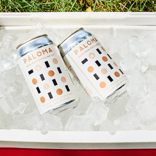 Two cans of Mean Mule Paloma drinks on ice in a cooler on grass.