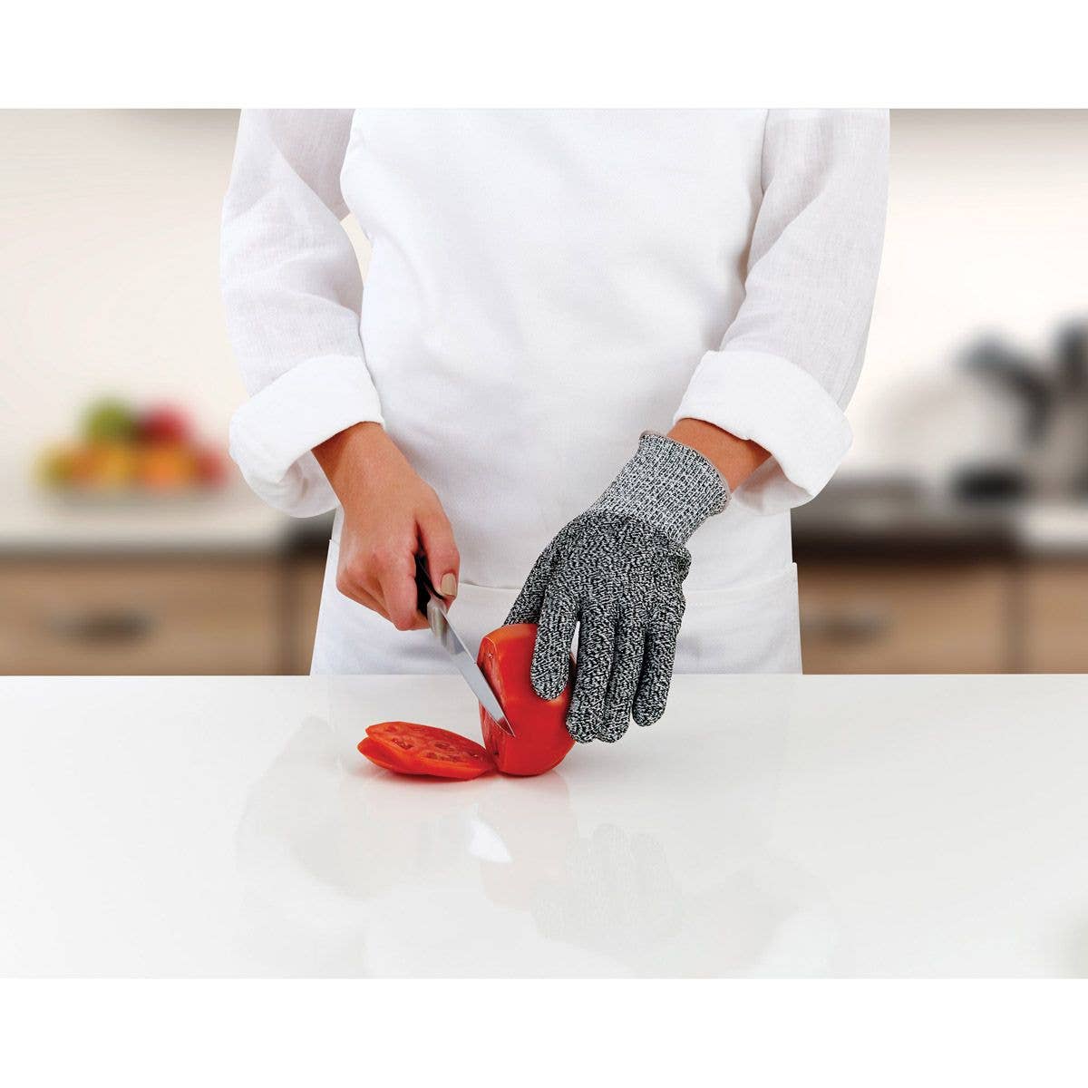 Person wearing a gray cutting glove while slicing a tomato on a white surface with a blurred kitchen background.