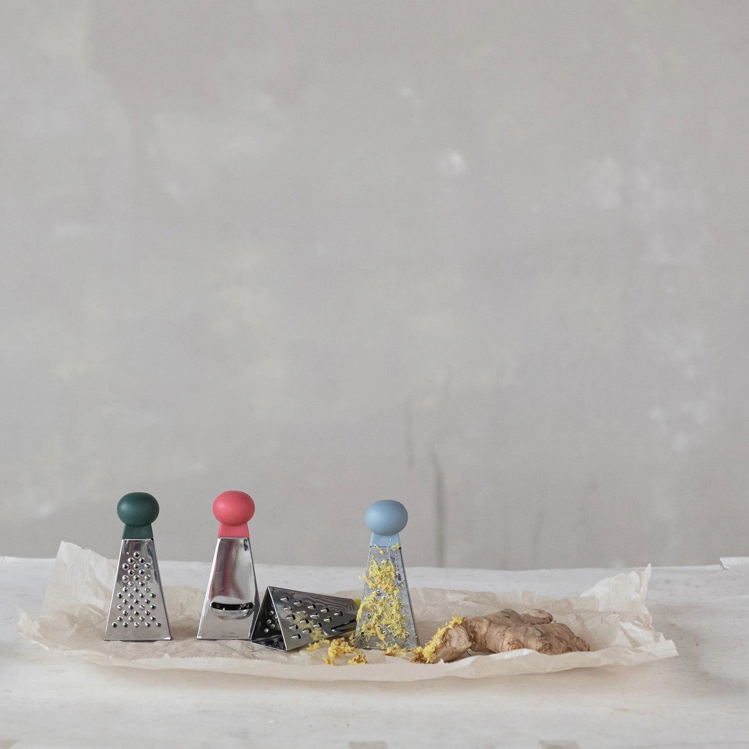 Four mini graters with colored handles, one lying on its side and another showing grated ginger with a piece of ginger lying nearby, all on a piece of paper on a neutral surface.