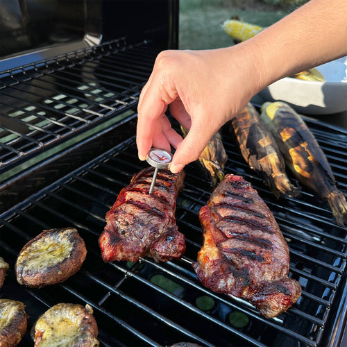 A person using a mini thermometer in meat outside on a grill.